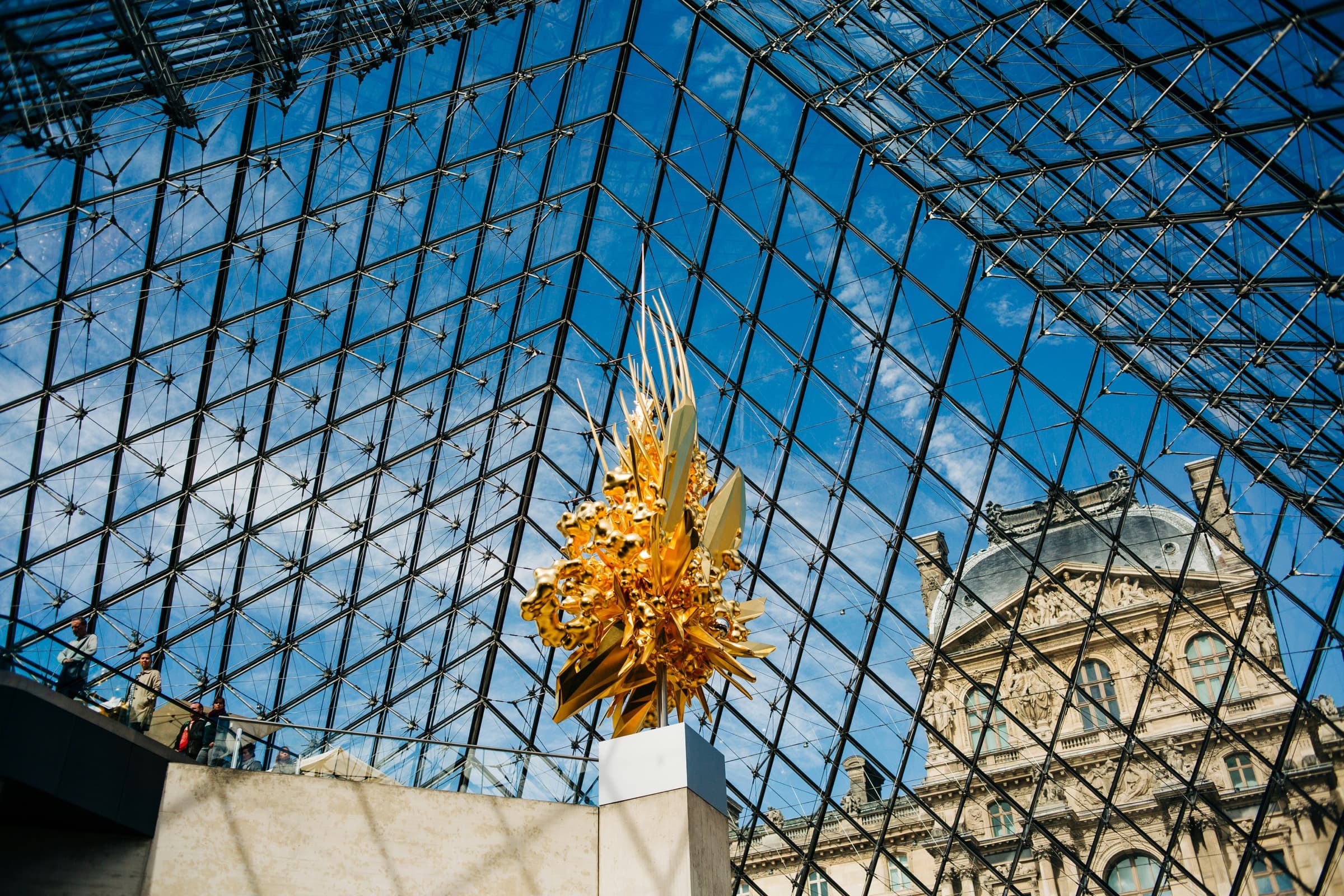 Golden sculpture inside the Louvre glass pyramid with blue sky and classical building visible through the geometric structure