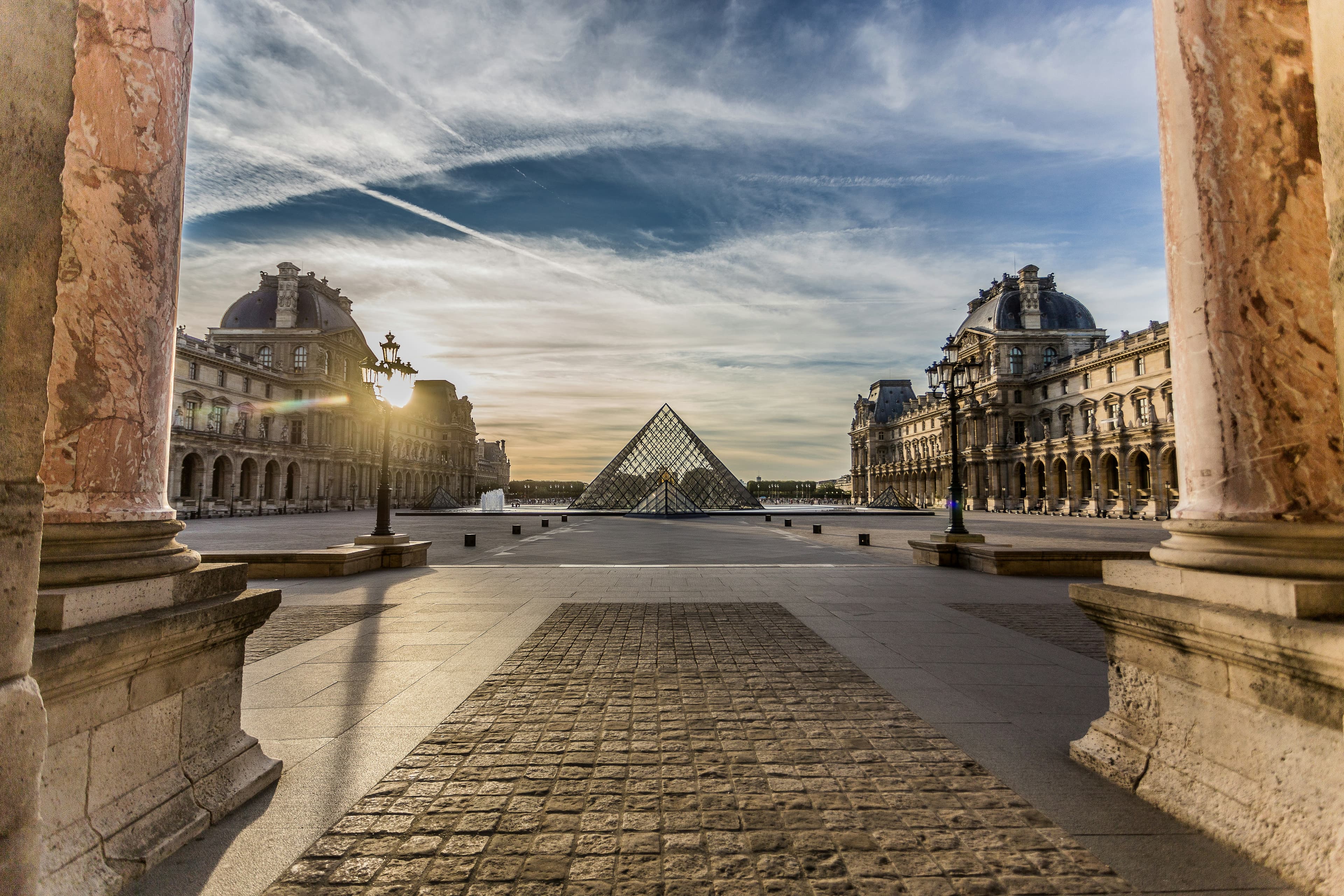 The Louvre Pyramid at sunset, framed between stone columns of the courtyard, long shadows converging on the glass structure