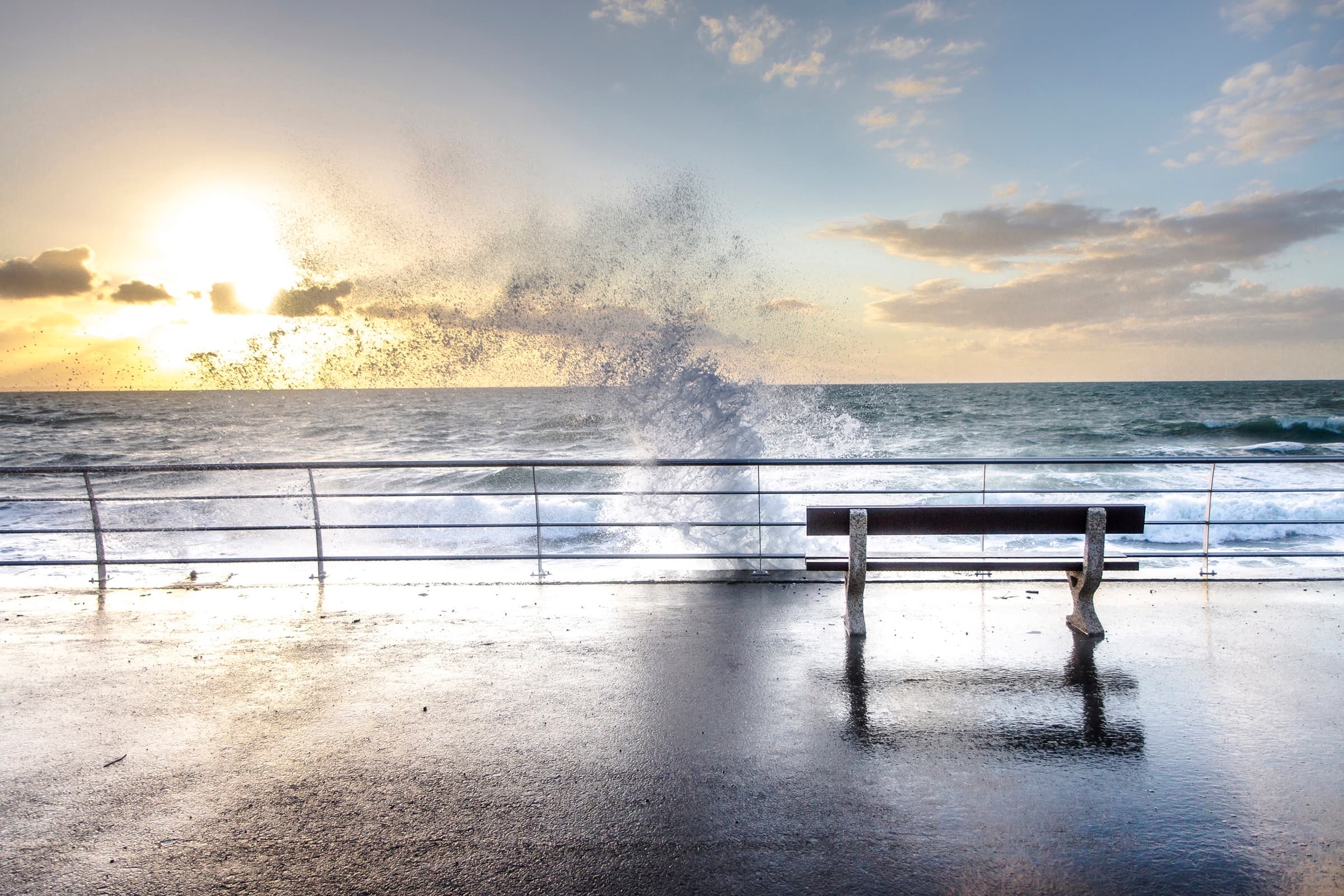 Ocean wave crashing over promenade bench at sunset